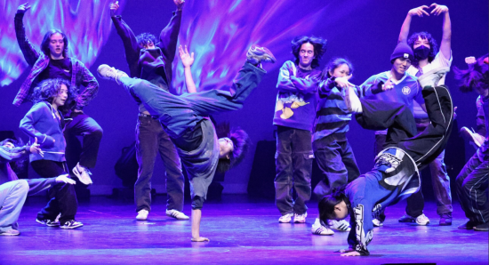 Image featuring breakers performing on stage. In the background are people cheering. In the foreground are 2 people in dynamic poses.
